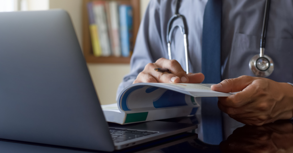 Surgeon sitting at desk in front of computer reading a textbook (model)